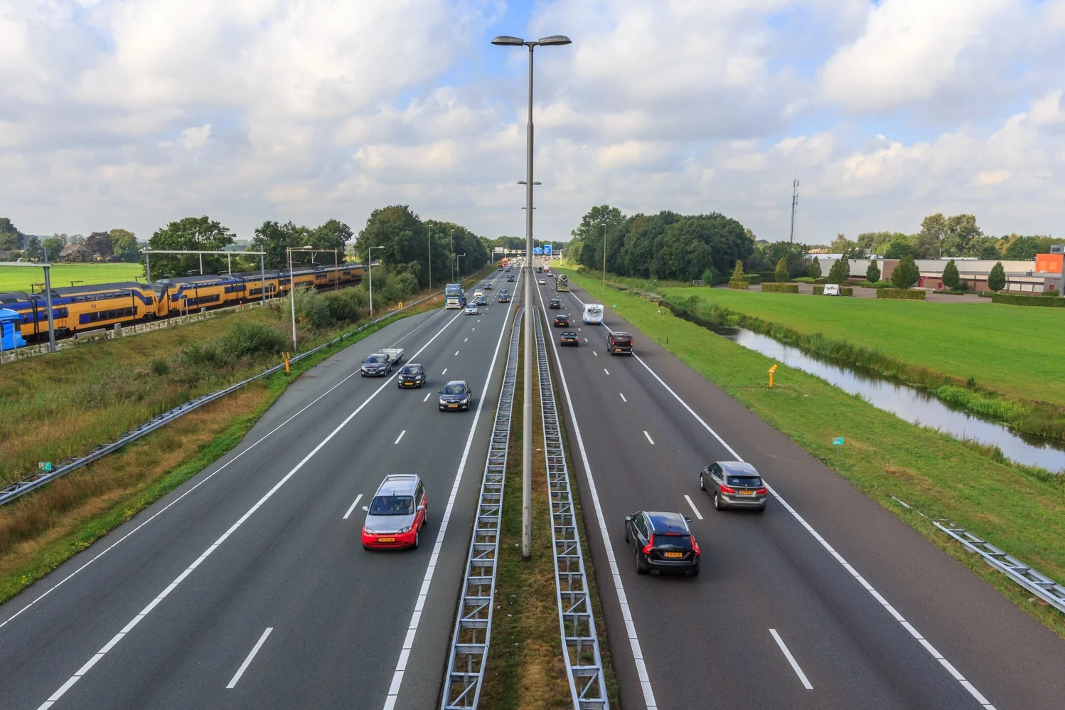 Drukke snelweg met naastgelegen spoorlijn waar een NS-trein rijdt door een groen, landelijk gebied.